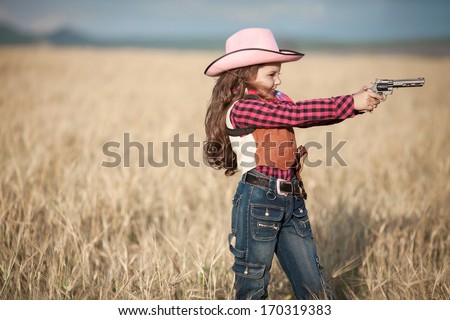 Cute Child having fun outdoors, little girl cowboy playing in wheat field at sunset. Happy baby girl with toy gun and cowboy hat enjoying nature. American Cowgirl. lovely smiling toddler portrait.