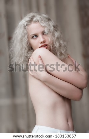 Half body portrait of topless young woman with long curly blond hair covering breasts with hands.
