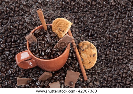 Coffee mug full of coffee beans on pile of coffee beans with chocolate and cinnamon sticks