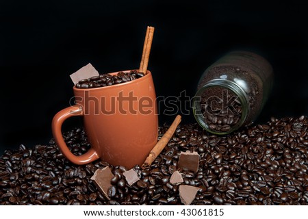 coffee mug full of coffee beans on pile of coffee beans with chocolate, cinnamon sticks and  jar of ground coffee