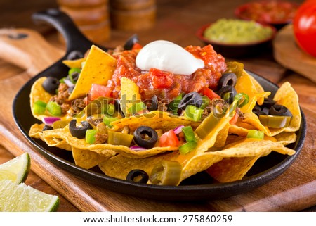 A plate of delicious tortilla nachos with melted cheese sauce, ground beef, jalapeno peppers, red onion, green onions, tomato, black olives, salsa, and sour cream with guacamole dip.