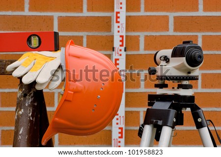 A leveling device, a hard hat, work gloves and a level against a wall shown in red bricks.