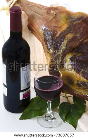 A front leg Serrano ham on displayed on a wooden rack and a bottle of red wine with a full glass of wine on a white background.
