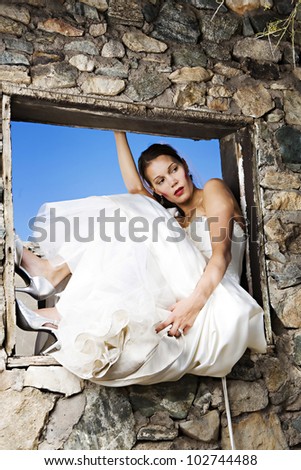A beautiful modern bride leans back in the window of an old building under a clear blue sky