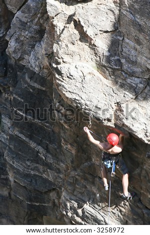 A rock climber works his way up a rock face protected by a rope clipped into bolts. He is wearing a helmet and quickdraws dangle from his harness. The route is in the desert southwest United States.