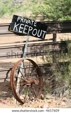 A sign stating \'PRIVATE KEEP OUT\' stands in front of a fence alongside an old wheel