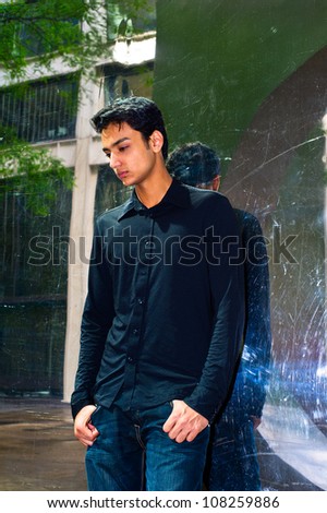 A young asian guy is standing against a mirror-like wall, lowering his head and thinking/Portrait of a young asian guy