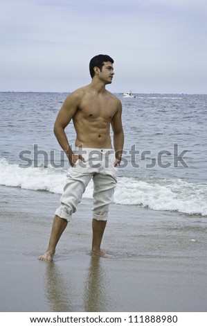 A young masculine guy is standing on the beach, facing waves of the ocean. A small boat on the background of ocean.
