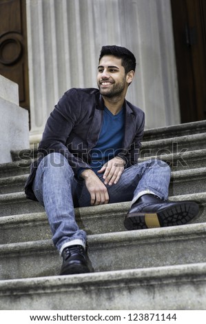 A young handsome guy is sitting on stairs, looking around and smiling./Big smile
