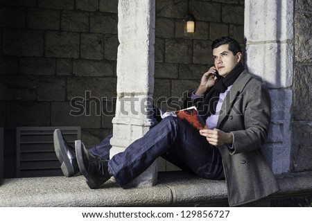A young guy is sitting on a old window frame,  reading and talking on the phone/Leisure Time