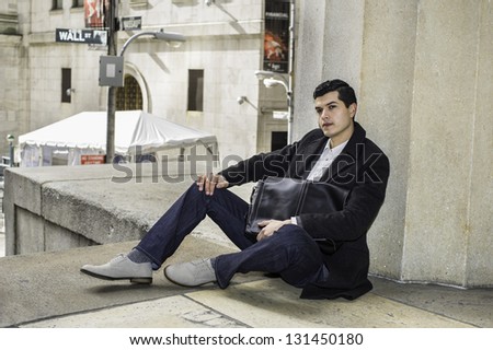 A young businessman hugging a bag  is sitting on the floor and take a break. There is a Wall Street sign on the background.