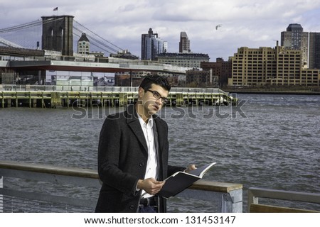 A young handsome scholar  is holding a book and standing on a river bank, looking around surrounding environment and thinking/Study Outside