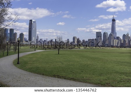 Looking faraway, the left side is Jersey City and the right side is New York City. The front ground is a small road in a green park./Jersey City and New York  in Spring Season