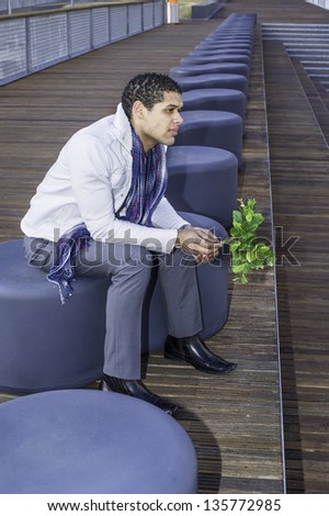 Holding a bunch of green leaves, a young guy is sitting on a modern style bench and waiting for you.