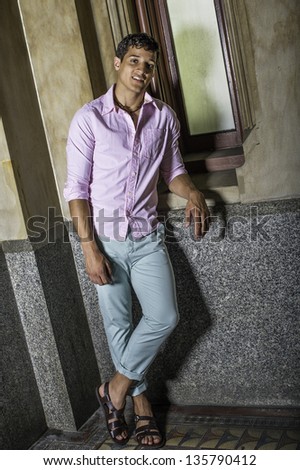 Dressing in a light pink shirt, light green pants and dark brown sandals, a young handsome guy is standing by a window and smiling./Portrait of Young Guy