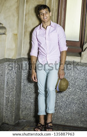 Dressing in a light pink shirt, light green pants and dark brown sandals,  a young handsome guy is standing by a window and holding a hat./Taking Break