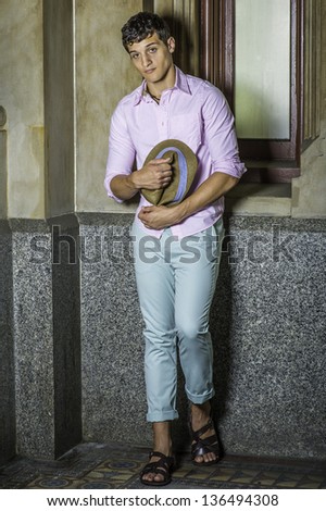 Dressing in a light pink shirt, light green pants and dark brown sandals,  a young handsome guy is standing by a window and holding his hat on his stomach./Taking Break