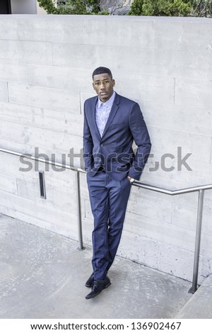 A young handsome black guy is standing outside against a wall, hands in pockets and confidently looking up./Portrait of Young Black Guy