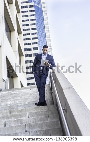 A young handsome black businessman is standing outside of a business building and reading at a small computer./Study Outside