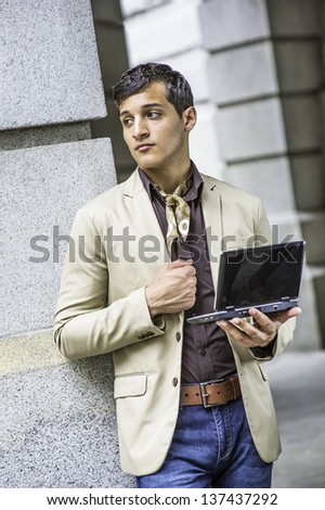 Dressing in a beige color blazer, a coffee color shirt, blue jeans, a scarf tied in the neck,  and holding a small computer,  a handsome young guy is confidently looking forward./Study Outside