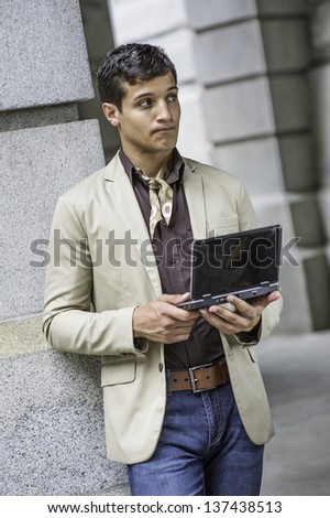 Dressing in a beige color blazer, a coffee color shirt, blue jeans, a scarf tied in the neck,  and holding a small computer,  a handsome young guy is closing his lips and thinking./Study Outside