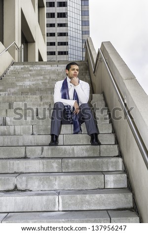 One hand touching his chin, a young student is sitting on steps outside a business building and into deeply thinking.