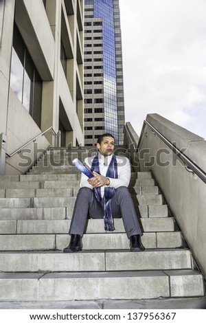 holding a magazine, a young student is sitting on steps outside a business building and into deeply thinking.