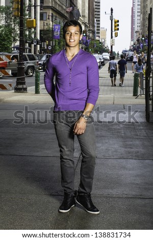 Wearing a purple sweater, a necklace,  black pants and black shoes, a young handsome guy is fashionably standing in a gateway to a busy street./Portrait of Young Guy