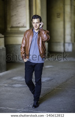 Dressing in a brown leather jacket, black pants and one hand putting in a pocket, a young handsome guy is confidently walking in a hallway and talking on the phone./Talking on Phone