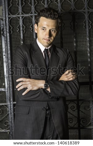 Crossing arms and biting lips a young businessman is seriously thinking outside a black metal gate./Deeply Thinking