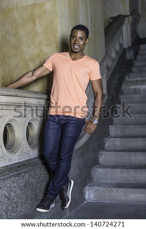 A young handsome black guy is standing by a staircase and smilingly looking at you./Portrait of Young Black Guy