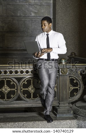 Dressing in white shirt, a black tie, gray pants, a young handsome black businessman is standing by a railing and working on a computer./Working Anywhere