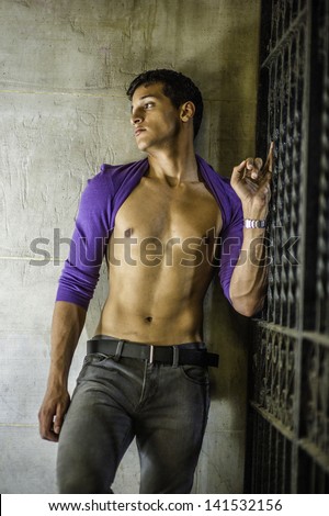 Standing on the corner between a wall and a metal gate, a sexy young guy is  rolling his purple sweater over his head, showing his well defined body, and into deeply thinking,/Summer Heat