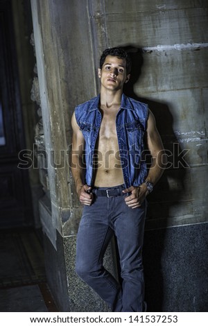 Dressing in a blue sleeveless Denim jacket, unbuttoned, a black pants , and standing in the front of a wall corner, a sexy young worker is taking a break outside./Take a Break