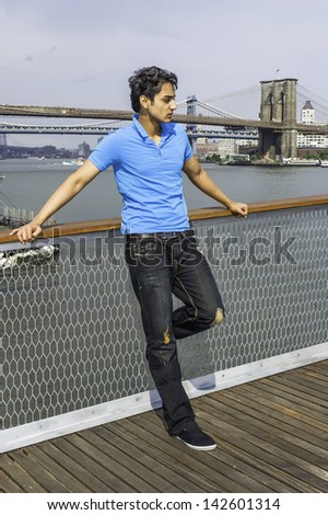 Dressing in a blue T shirt, fashionable pant, standing on the dock, a young attractive asian teenager is looking down and into deeply thinking. The background is a big bridge in a urban scene.