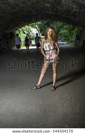 A pretty woman with long brown curly hair, dressing in a skirt,  is  standing under the shade in summer time,  thoughtfully looking forward./Under the Shade