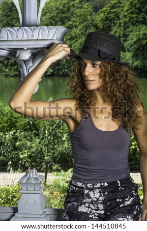 Dressing in a gray tank and wearing a sun hat, a pretty woman with long brown curly hair is  standing in a park and into deeply thinking./Pretty Woman in Summer