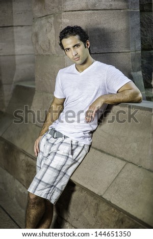 Dressing in white T shirt and pattern pants, a young handsome guy with bright piercing eyes is leaning on a rocky wall, relaxing and thinking./Relaxing and Thinking