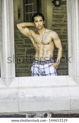 Half naked and wearing a cross necklace, a handsome, muscular guy is standing by an ancient style window frame and looking outside./Portrait of Young Fitness Guy