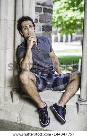 Dressing in a gray polo shirt, pattern shorts and black leather sneakers, one hand touching his chin,  a young strong handsome guy is sitting on a old fashion window frame and into deeply thinking.