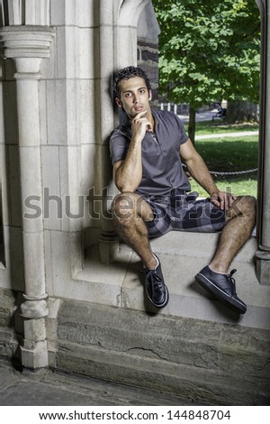 Dressing in a gray polo shirt, pattern shorts and black leather sneakers, one hand touching his chin,  a young strong handsome guy is sitting on a old fashion window frame and looking at you.