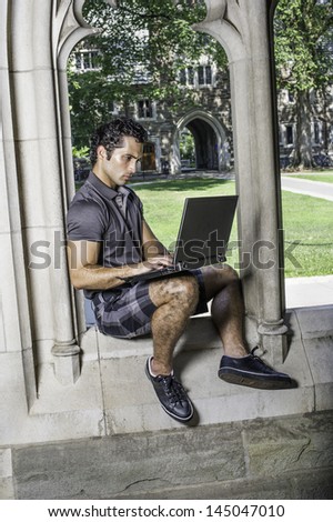 Dressing in a gray polo shirt, pattern shorts and black leather sneakers, a young handsome student is sitting on a old fashion window frame and study on a computer in a university campus
