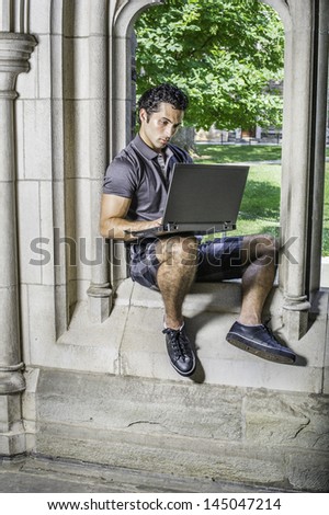 Dressing in a gray polo shirt, pattern shorts and black leather sneakers, a young handsome student is sitting on a old fashion window frame and study on a computer in a university campus