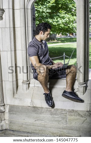 Dressing in a gray polo shirt, pattern shorts and black leather sneakers, a young handsome student is sitting on a old fashion window frame and study on a computer in a campus /Study by a window
