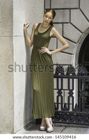 Dressing in a green Maxi Tank Dress, high heal shoes, a young fashion black girl is standing  by a pattern wall and a black metal railing and smilingly looking at you./Portrait of Young Fashion Girl
