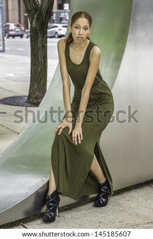 Dressing in a green Maxi Tank Dress, a young black girl is sitting on a metal structure in a street and relaxing. /Relaxing Outside