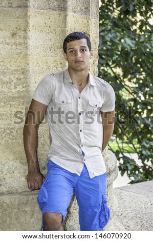 Dressing in a iron short sleeve shirt, blue shorts, a young attractive guy is standing by a column and looking at you. /Portrait of Young Guy