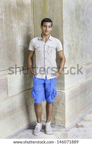 Dressing in a iron short sleeve shirt, blue shorts, a young attractive guy is standing in the corner of walls and looking at you. /Portrait of Young Guy