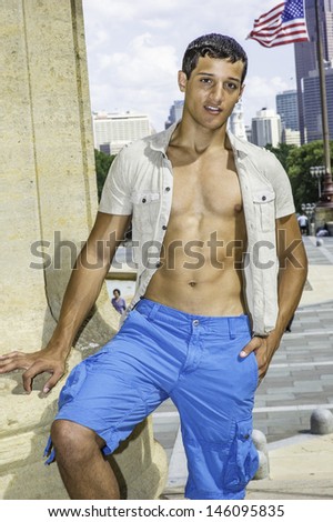 Dressing in short sleeve shirt, unbuttoned, and blue shorts, a young attractive guy is charmingly looking at you. /Portrait of Young Guy