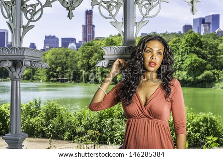 Dressing in a red dress, a pretty black girl with long curly hair is standing by a pavilion in the park, thoughtfully looking forward. / Thinking about You